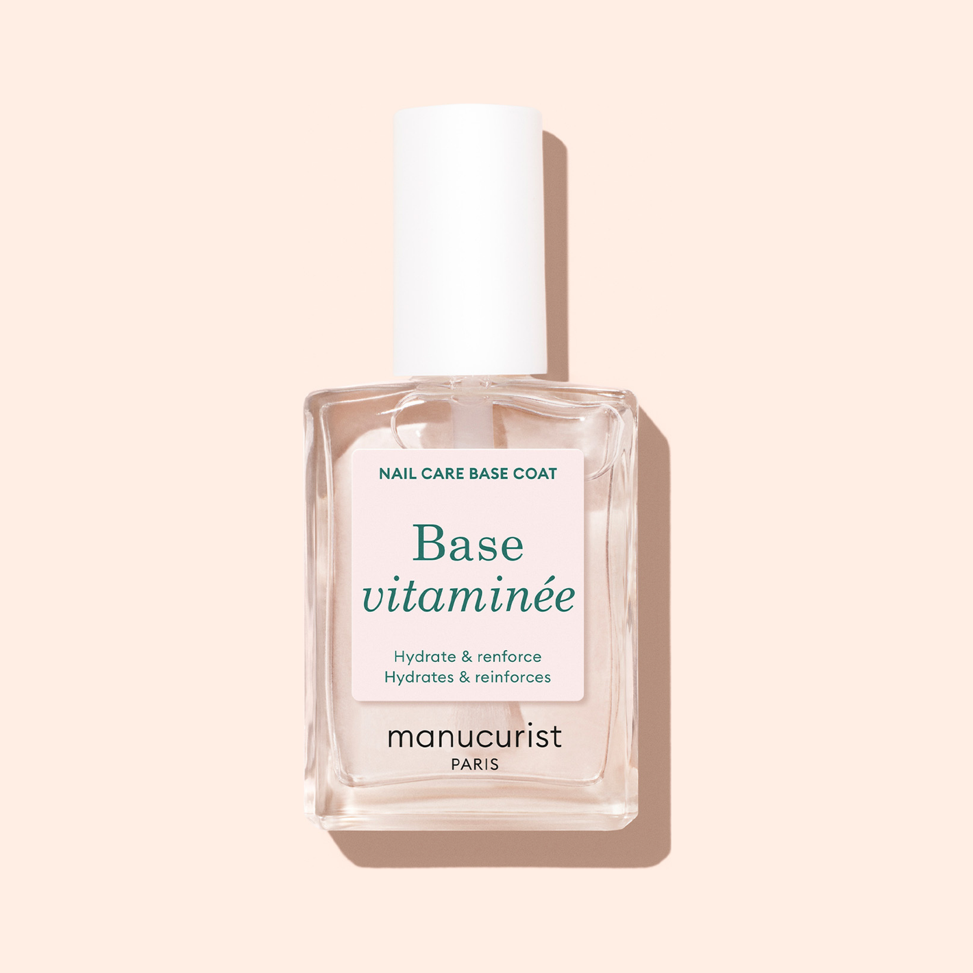 A clear glass bottle of Manucurist Vitaminised Base with a white cap sits on a light beige background. The pale pink and white label, with blue and black text, highlights this nail-protecting and moisturizing polish.