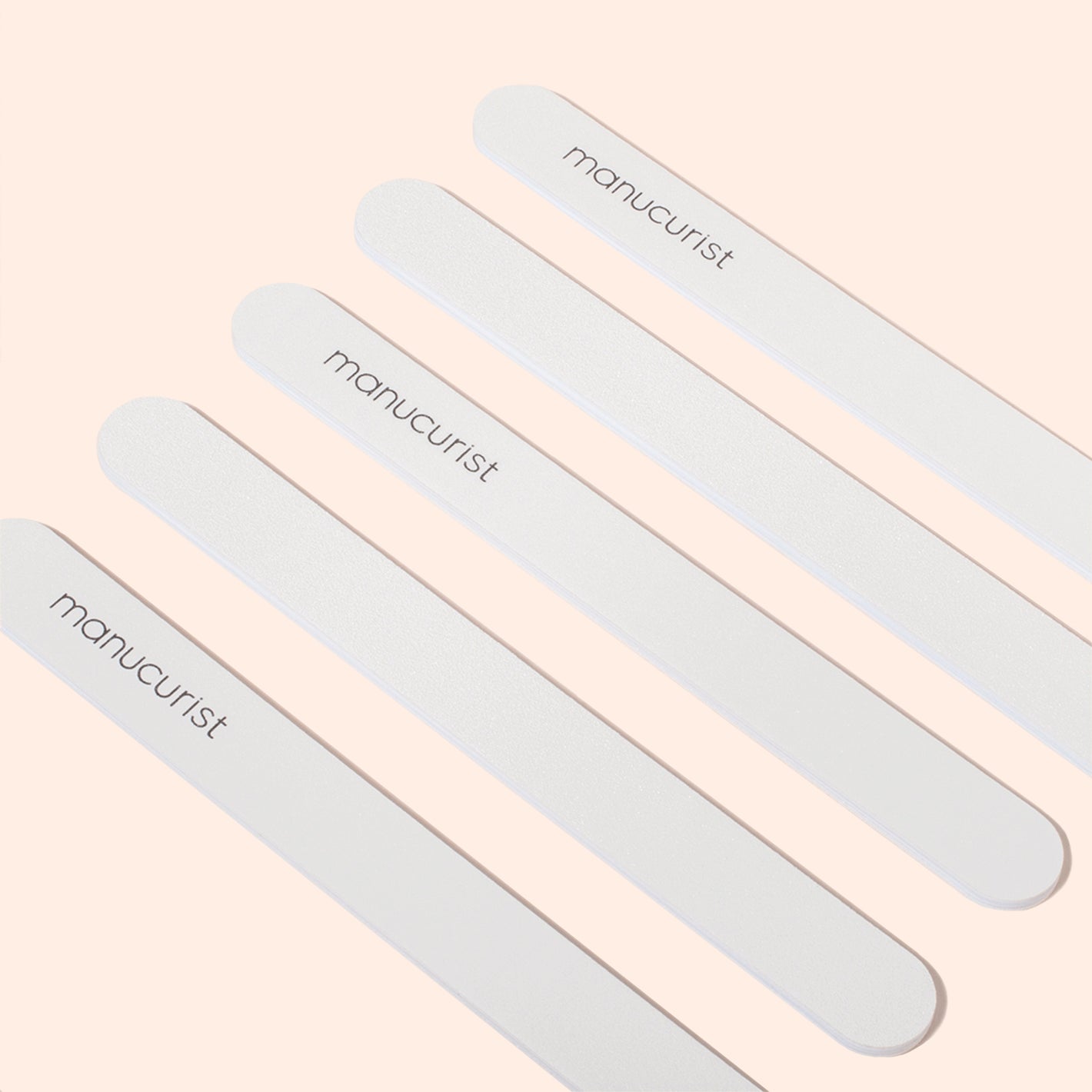 Five white emery boards with rounded ends, each labeled Manucurist, are arranged diagonally on a pale peach background—perfect for gently smoothing delicate nails.