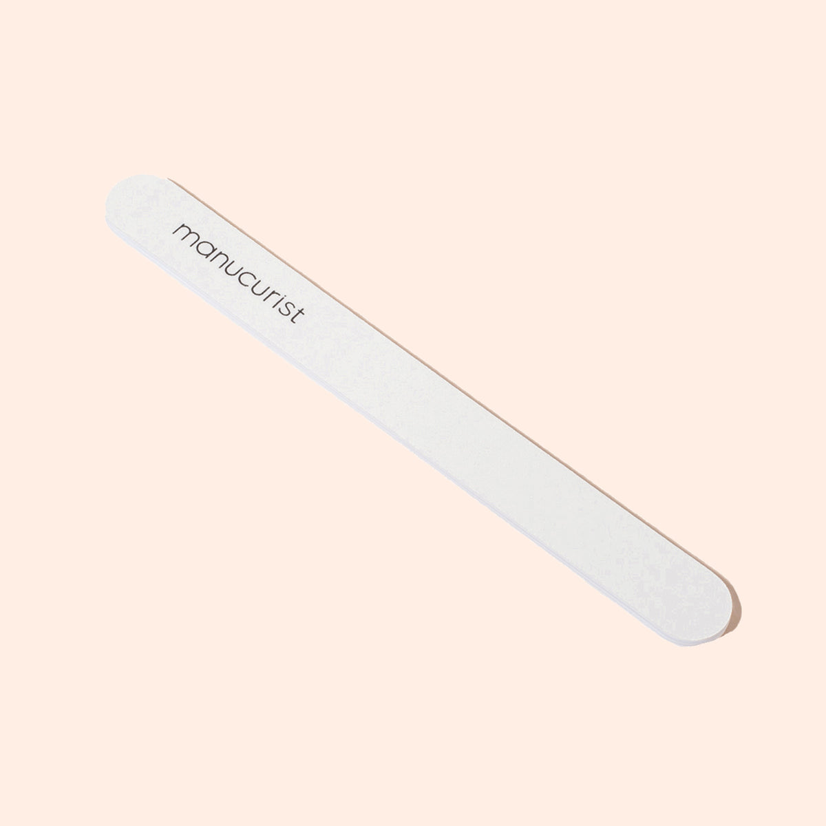 A Manucurist 100/180 emery board for natural nails with rounded edges and the brand name printed on it, featuring soft and coarse grits, displayed on a pale peach background.