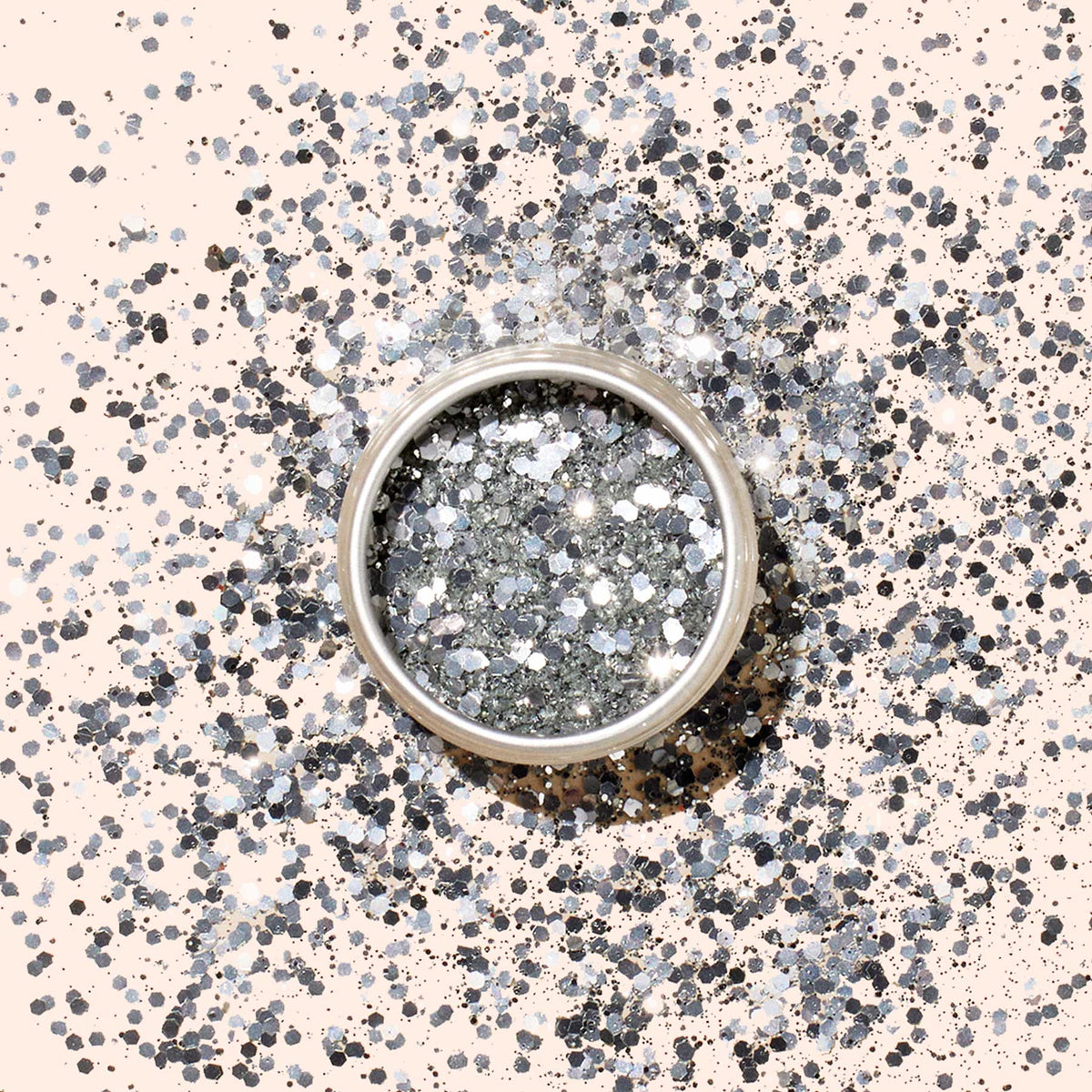 A round container of Manucurist Glitter Duo Silver  Mirror sits on a light beige surface, surrounded by scattered silver glitter and sparkling dots that highlight its pearlescent shine.