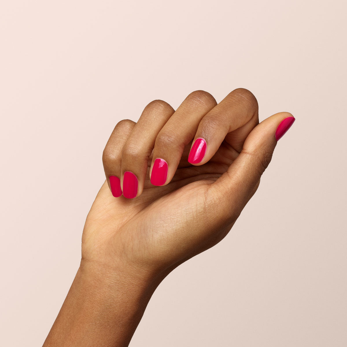 A hand with medium brown skin tone is shown against a light beige background. The nails are neatly manicured and painted with Manucurist-test’s Peonie OLD, a bright pink vegan nail polish. The fingers are relaxed and slightly curled inward.