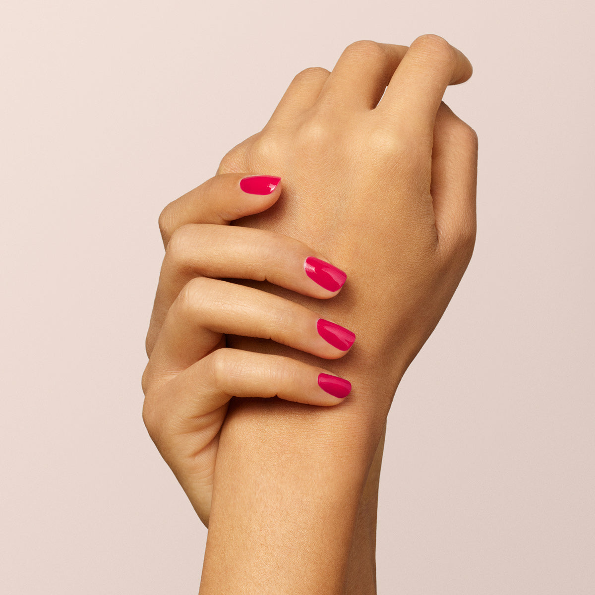 A hand with neatly manicured nails painted bright pink using Manucurist-test’s Peonie OLD vegan nail polish gently holds the wrist of the other hand, against a light beige background.