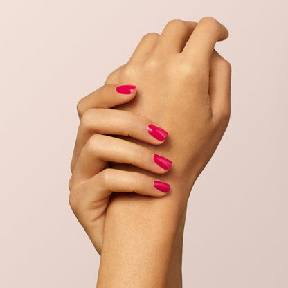 A hand with neatly manicured nails painted bright pink using Manucurist-test’s Peonie OLD vegan nail polish gently holds the wrist of the other hand, against a light beige background.