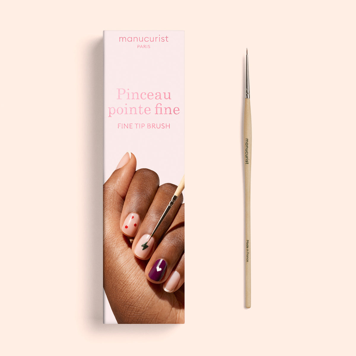 A Fine Tip Brush with vegan synthetic bristles rests next to its packaging, which showcases a hand adorned with assorted nail art designs. The box is labeled Manucurist and Fine Tip Brush, emphasizing its suitability for detailed and precise nail art.