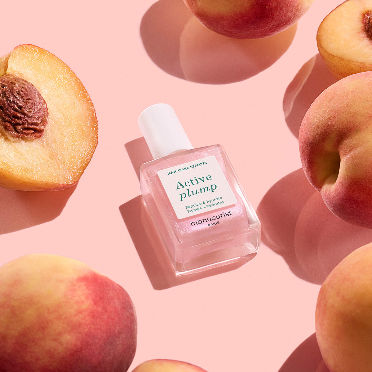 A bottle of Manucurist Active™ Plump Pink Glazed, a plant-based nail care essential, sits on a pink surface, surrounded by whole and halved peaches.