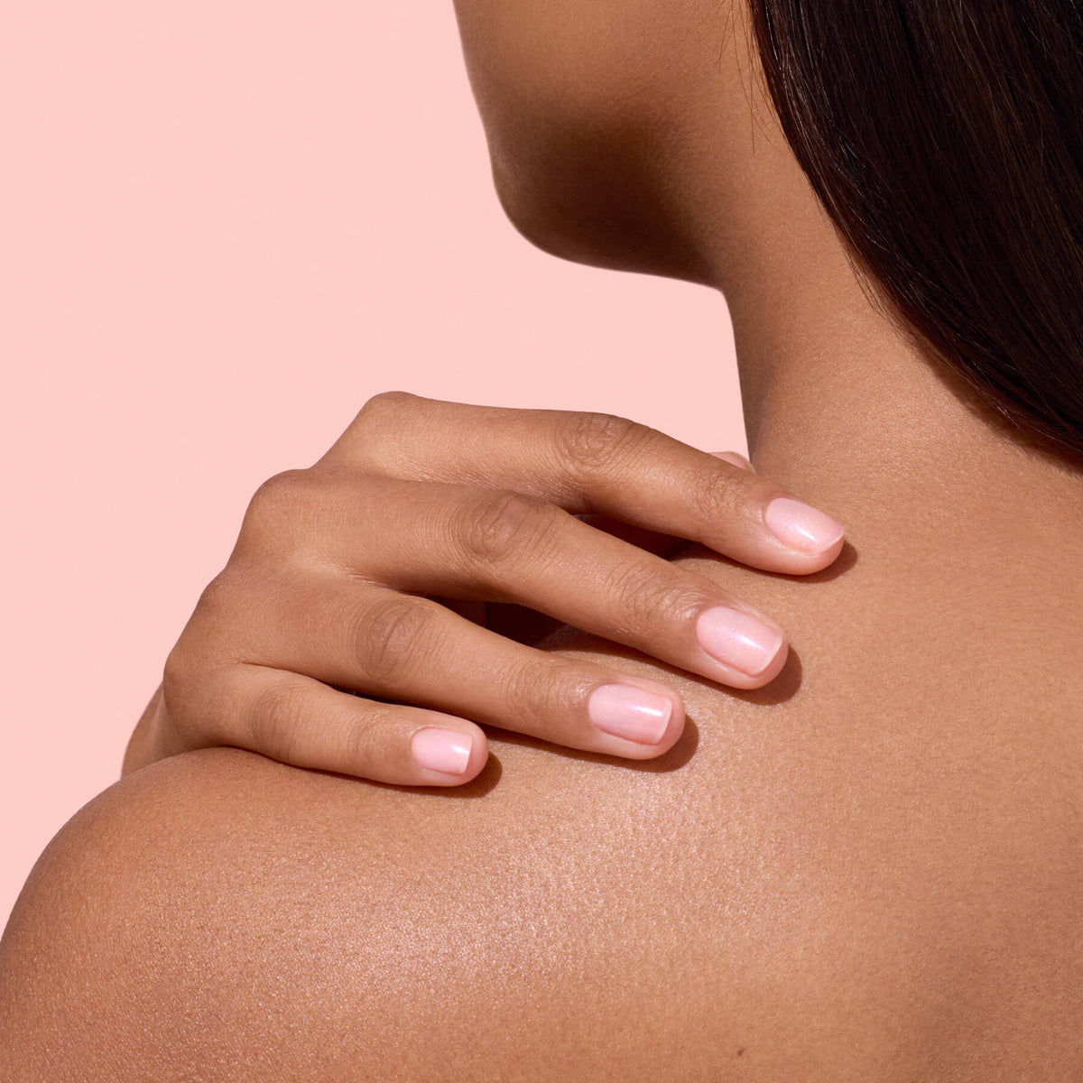 A person with medium skin tone rests their manicured hand, showcasing hydrated nails polished in Manucurist’s Active™ Plump Pink Glazed, on their bare shoulder against a soft pink background.