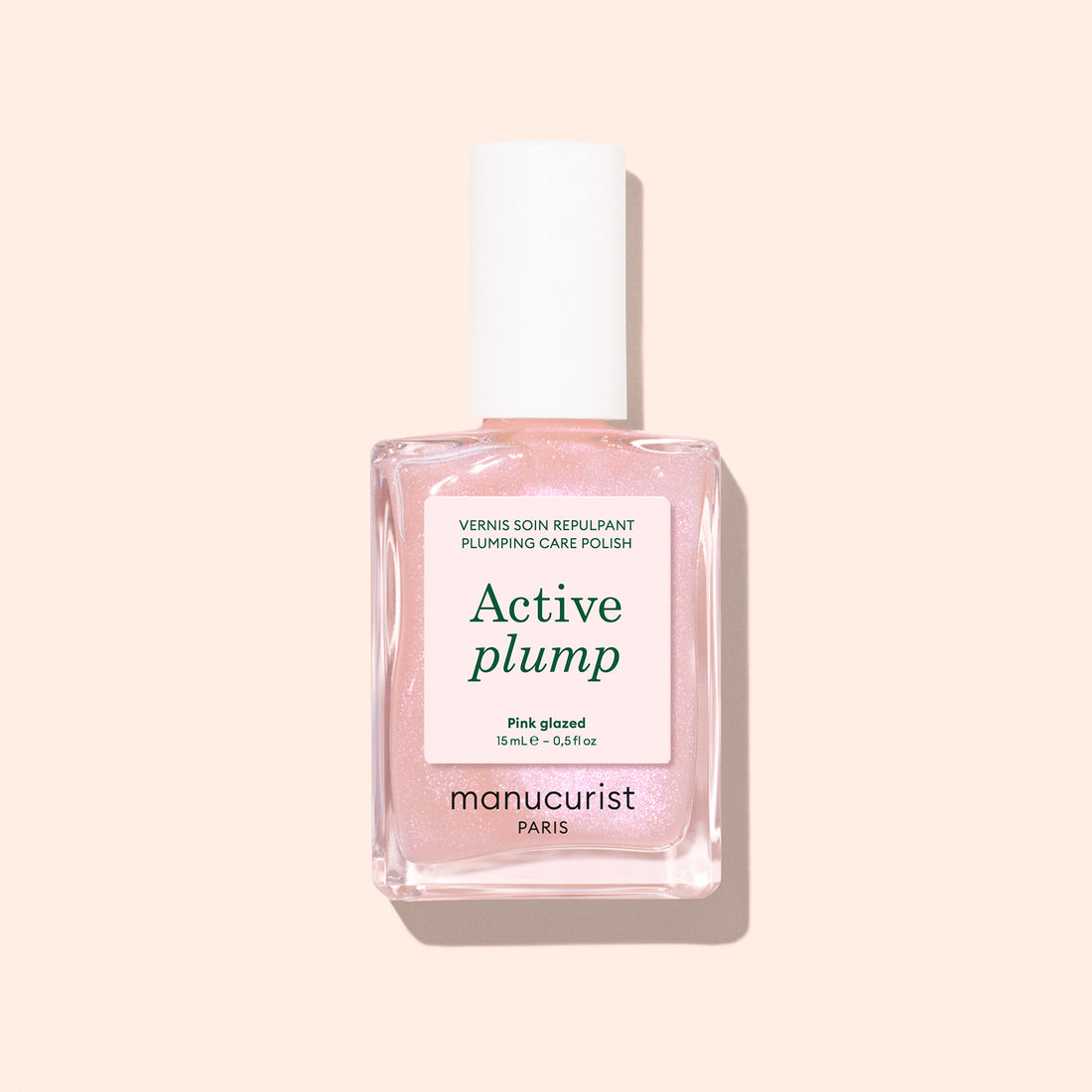 A bottle of Manucurist Active Plump Pink Glazed nail polish with a white cap and light pink formula sits on a pale peach background, its label showcasing plumping and hydrating benefits for beautifully moisturized nails.