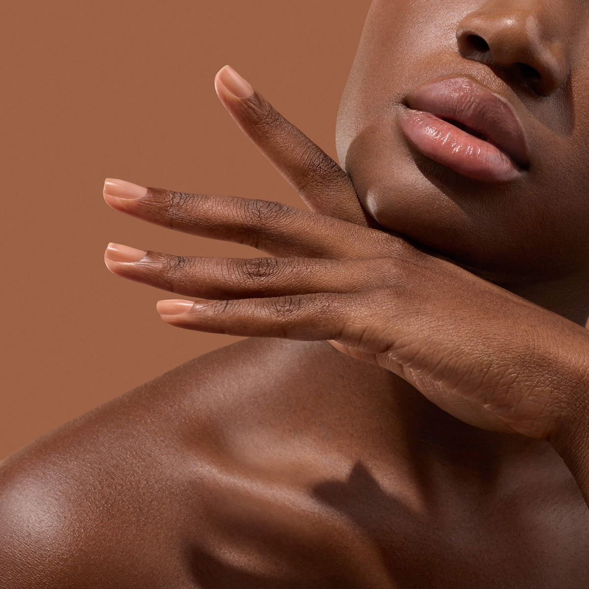 A close-up of a woman’s face and hand against a brown background, showcasing smooth skin, glossy lips, and neatly manicured nude nails treated with Manucurist Active™ Smooth 03 for the ultimate in nail care.