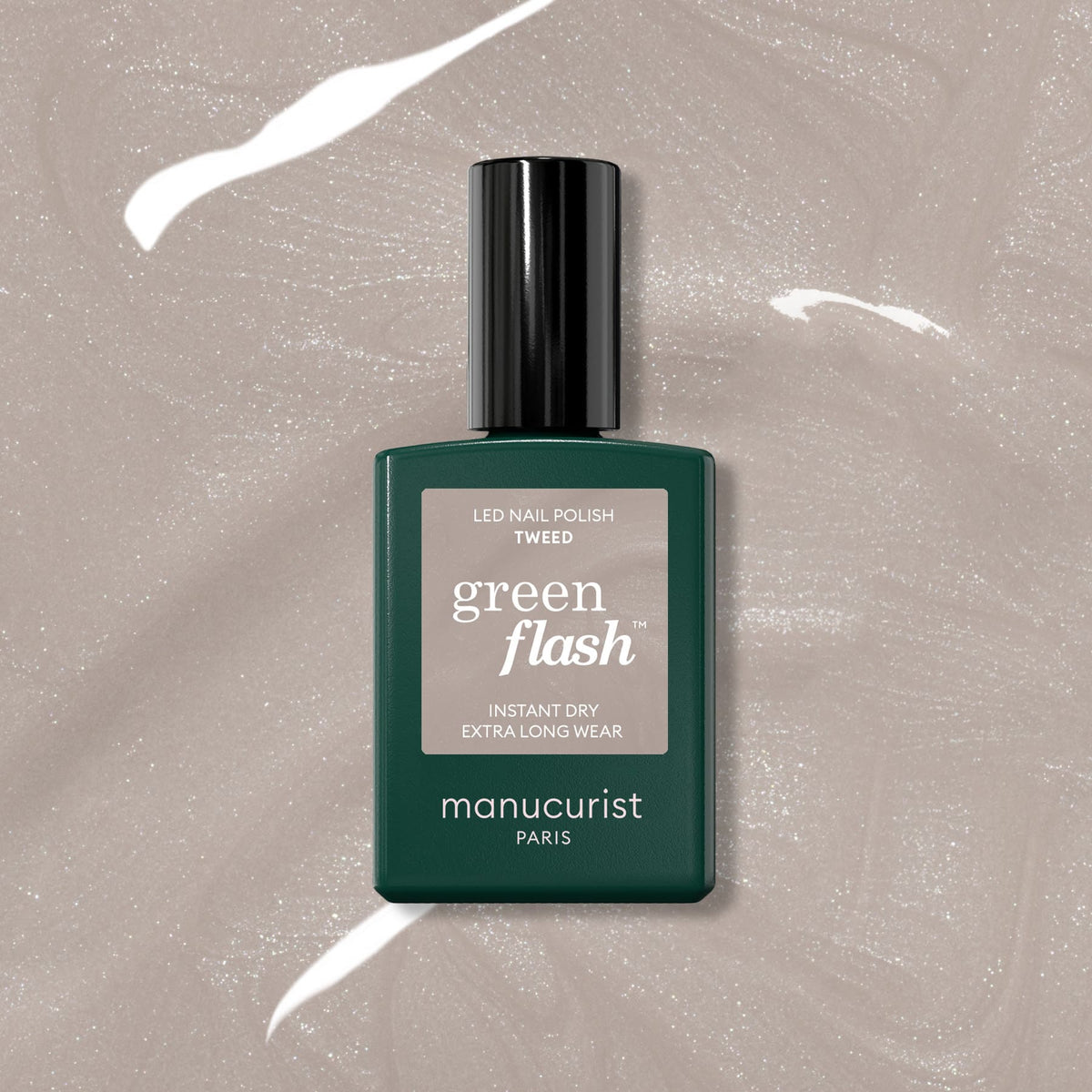 A bottle of Manucurist Tweed LED Gel Polish sits on a shiny, beige surface. The dark green bottle with black cap and light label suggests the polish’s pearlescent, warm grey shade.