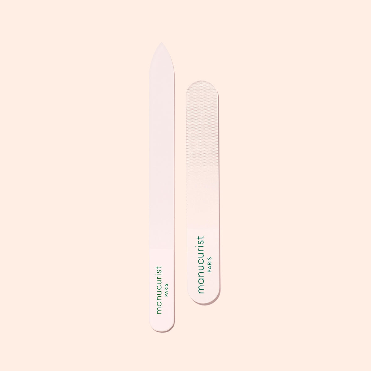 Two Manucurist Glass Nail Files with white surfaces and green logos rest side by side on a light beige background. One has a pointed tip and the other is rounded, both featuring smooth, premium designs.