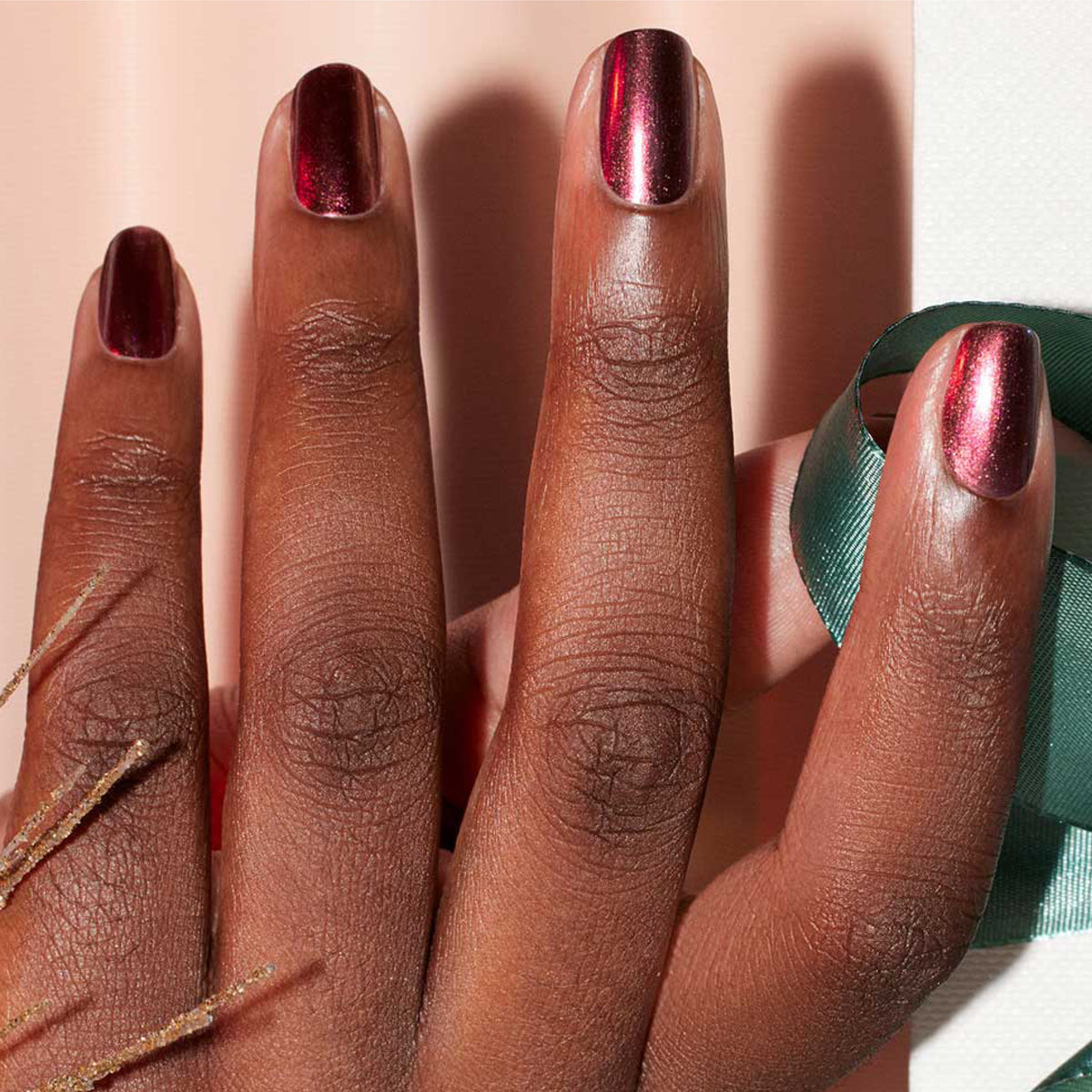 A close-up of a dark-skinned hand with short nails painted in Manucurist’s Utopia, a metallic crimson gel polish, holding a green ribbon against a light background with dried plant stems.