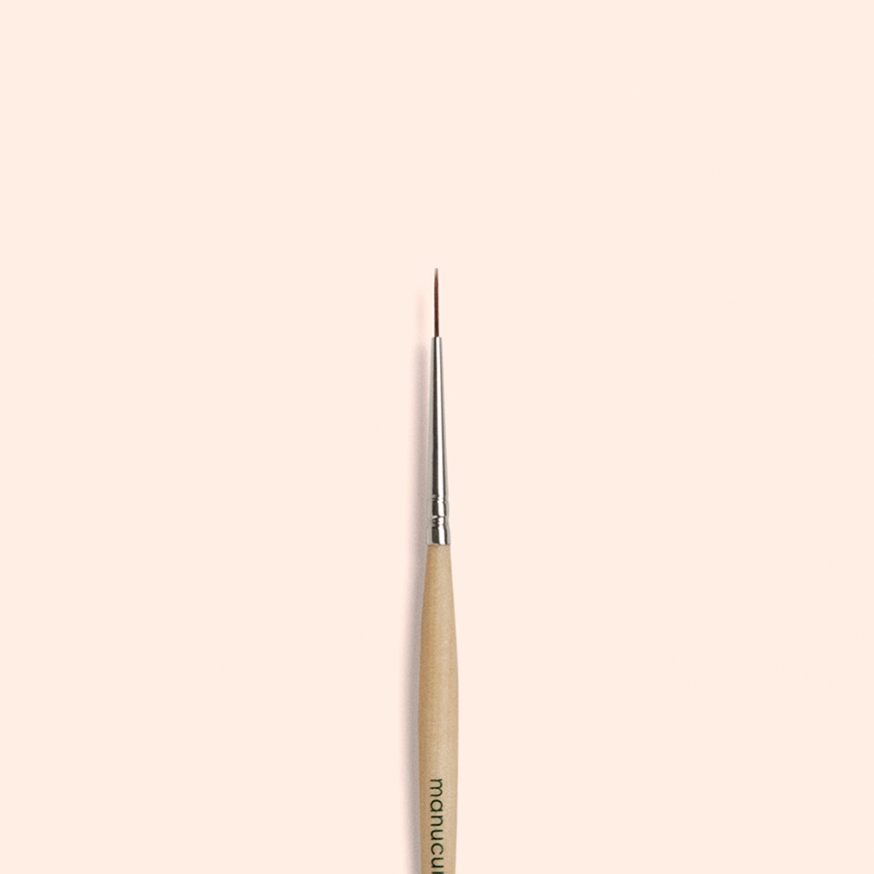 The Manucurist Liner Brush, featuring a thin, fine tip with a wooden handle and silver ferrule, is centered vertically on a plain beige background—perfect for creating precise nail art designs.