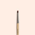The Manucurist Glitter Brush, featuring a wooden handle, metal ferrule, and rounded vegan bristles, stands vertically on a light beige background—ideal for creating radiant nail art.