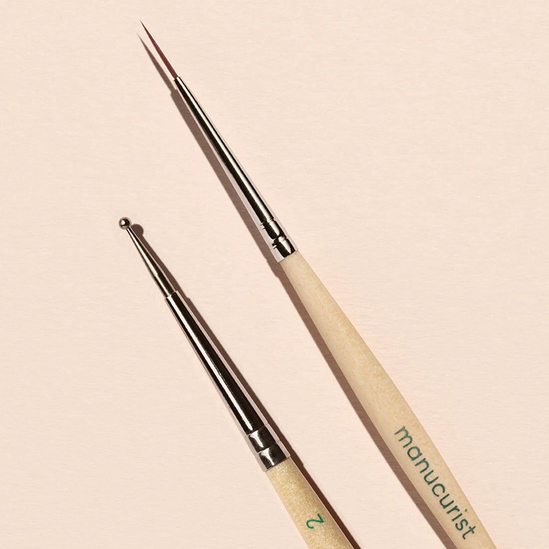 The Manucurist Dotting Tool Nail Art Liner Brush Duo features light wooden handles and silver ferrules resting on a beige background. One brush has a fine liner tip; the other is a small metal ball dotting tool.