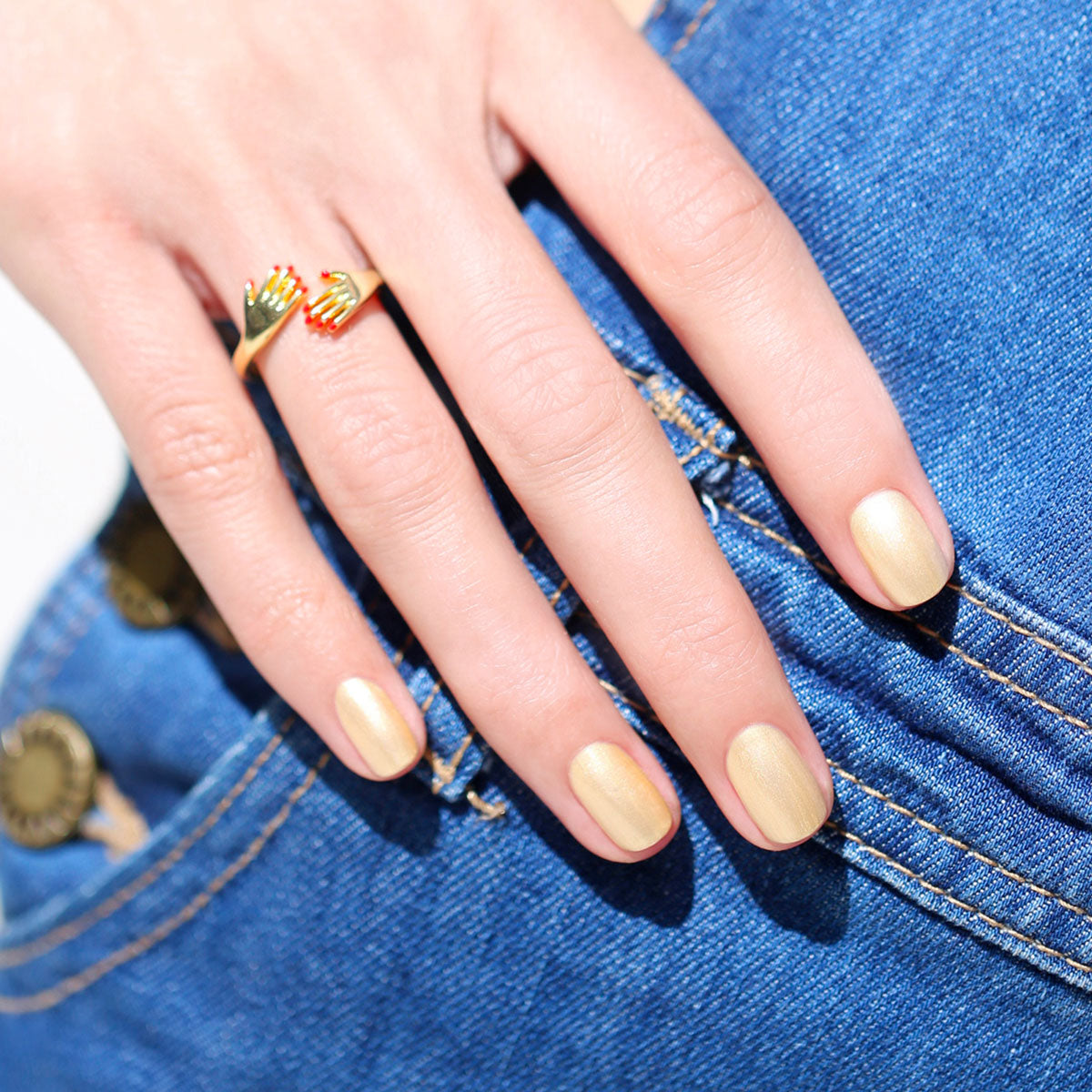 A hand adorned with Manucurist’s Gold Sand shimmering yellow metallic nail polish and a gold ring rests on blue denim jeans with visible stitching and a brass button.