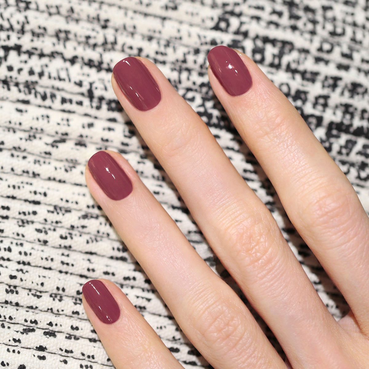 A hand with neatly manicured nails painted in Manucurist’s Victoria Plum Green Flash gel polish rests on a textured black and white patterned surface.