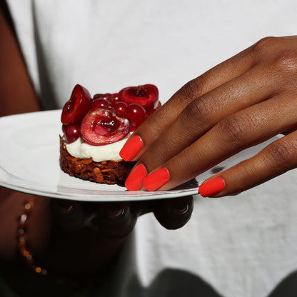 A hand adorned with Manucurist-test’s Red Coral OLD, a vibrant orange-red, long-lasting gel nail polish, holds a white plate topped with a cherry dessert. The person is wearing a white shirt and a bracelet.