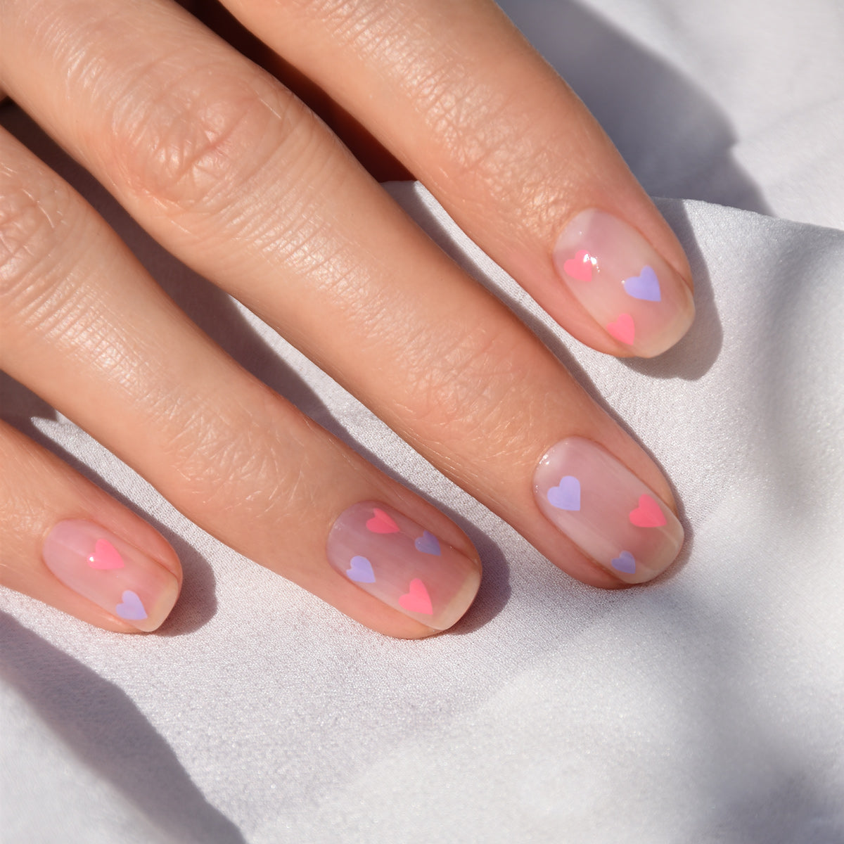 A hand with natural, clear nails decorated with tiny pink and purple heart designs—created using the Manucurist Dotting Tool  Nail Art Liner Brush Duo—rests on a white fabric background.