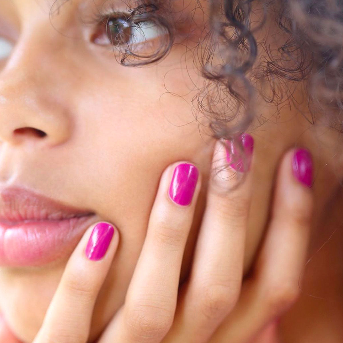 A close-up of a woman with curly hair touches her face, showcasing shiny pinky mauve "Armeria" nail polish by Manucurist on her fingernails. She has a calm, thoughtful expression and natural makeup.