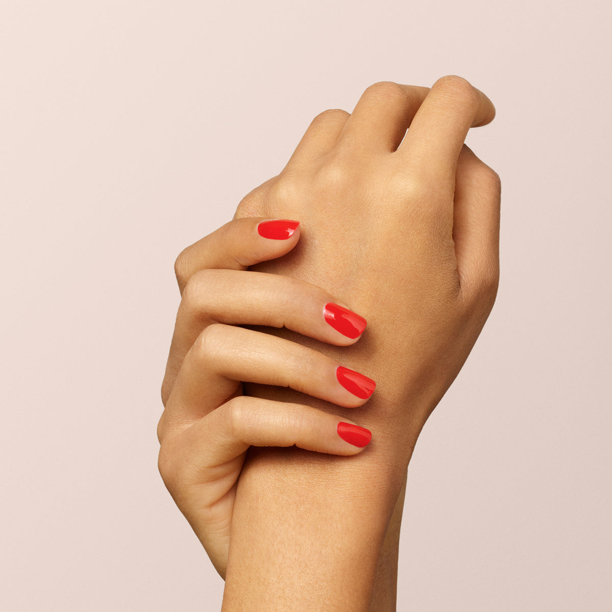Two hands with neatly manicured nails painted bright red using Manucurist-test’s Red Coral OLD gel nail polish are gently clasped together against a plain, light beige background.
