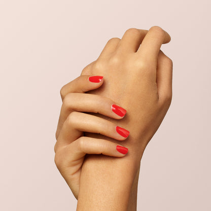 Two hands with neatly manicured nails painted bright red using Manucurist-test’s Red Coral OLD gel nail polish are gently clasped together against a plain, light beige background.