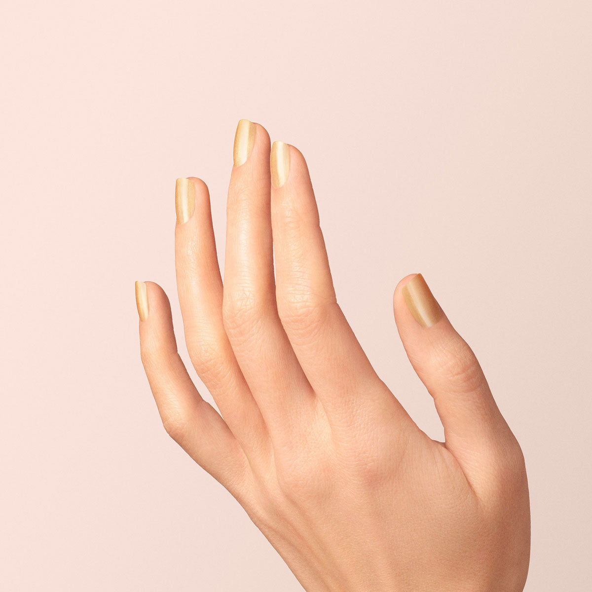 A hand with neatly manicured nails painted in Manucurist’s Gold Sand, a shiny, iridescent yellow nail polish, shown against a soft, light beige background.