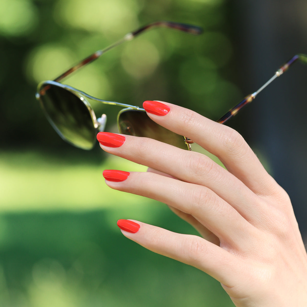 A hand with Manucurist-test Red Coral OLD gel nail polish holds a pair of sunglasses outdoors, set against a blurred green background.