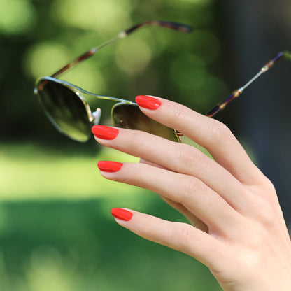 A hand with Manucurist-test Red Coral OLD gel nail polish holds a pair of sunglasses outdoors, set against a blurred green background.