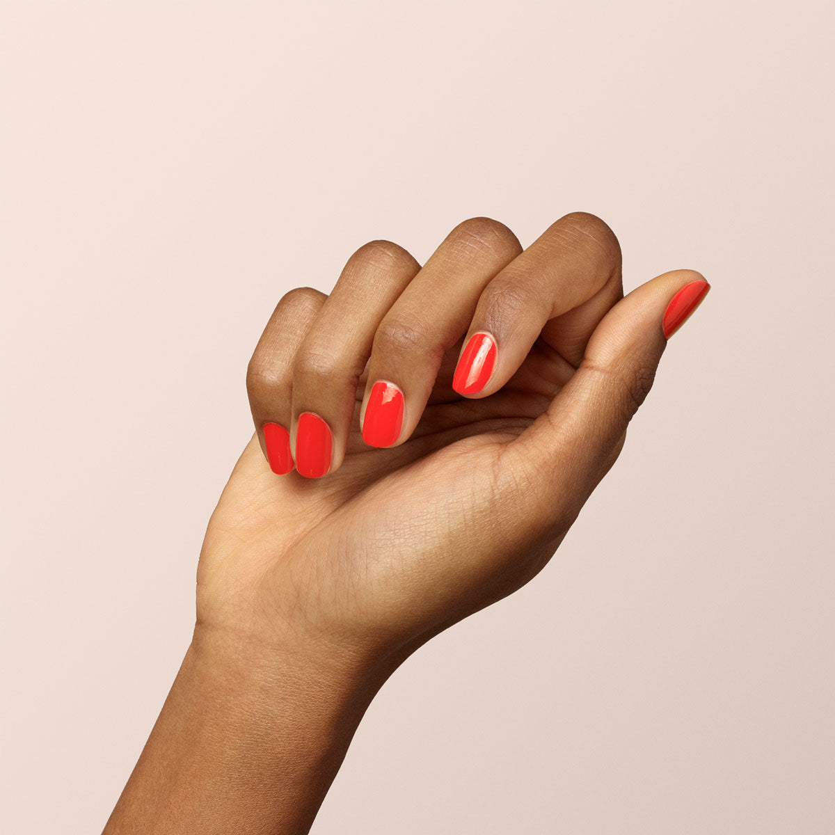 A hand with medium brown skin tone displays nails painted in Manucurist-test’s Red Coral OLD glossy gel nail polish against a plain, light background, showcasing a long-lasting manicure.