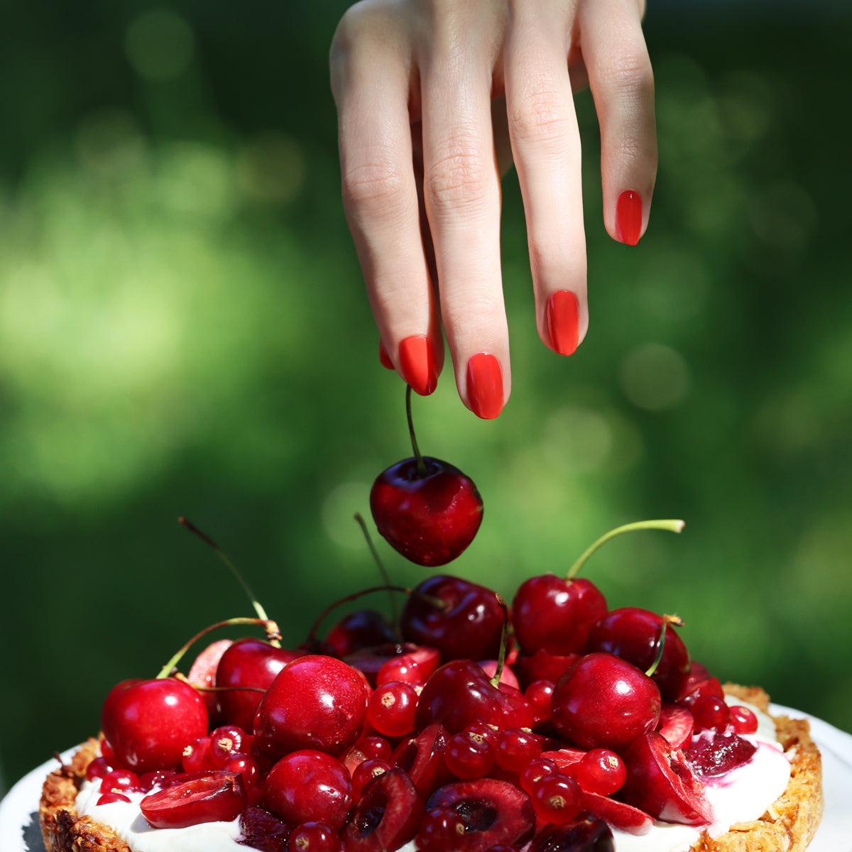 A hand with red-painted nails in Red Coral OLD by Manucurist-test holds a cherry above a dessert topped with fresh cherries and cream, set against a blurred green outdoor background.