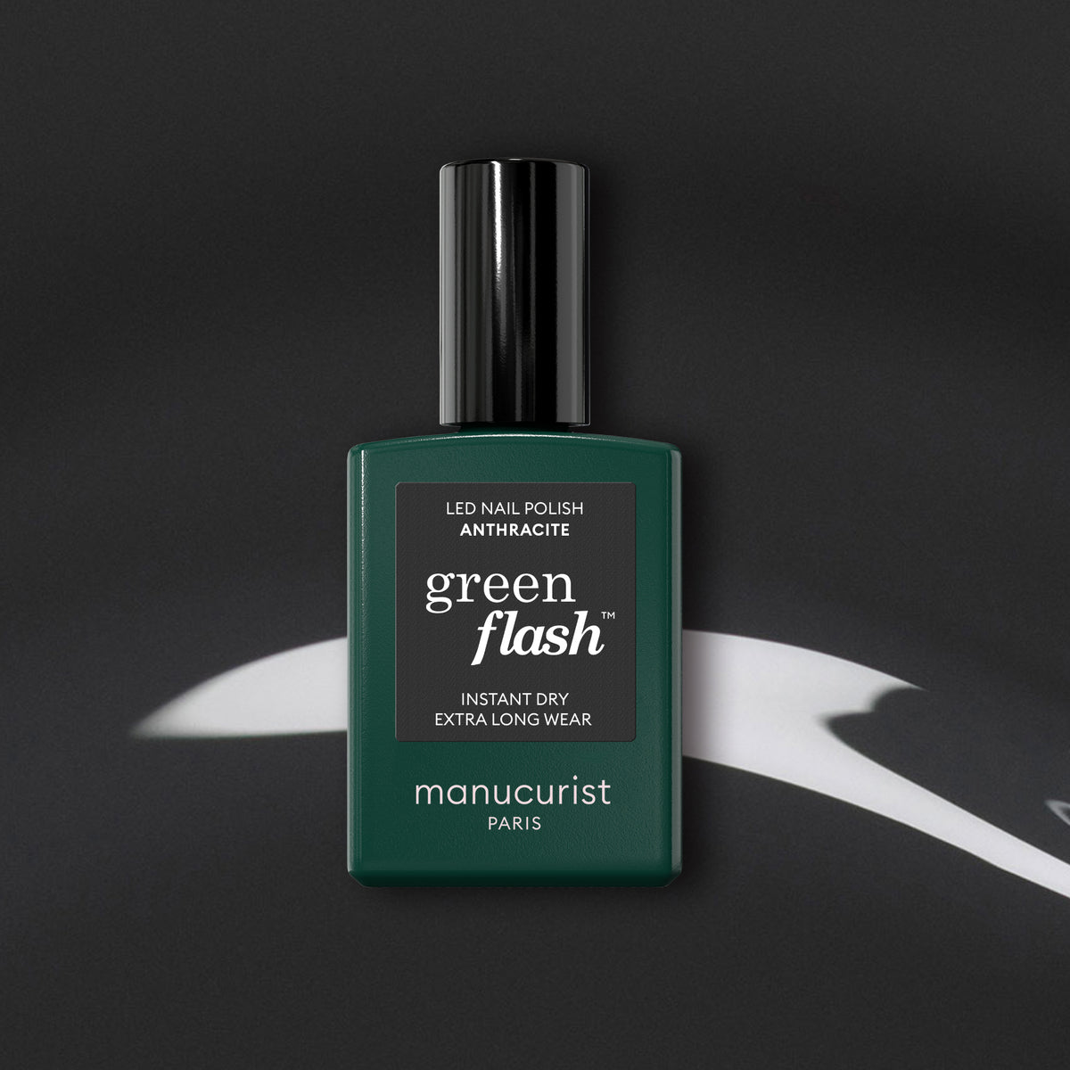 A dark green rectangular Manucurist bottle of Green Flash gel polish in the Anthracite shade, a chic dark grey, with a black cap sits on an abstract black and white background.