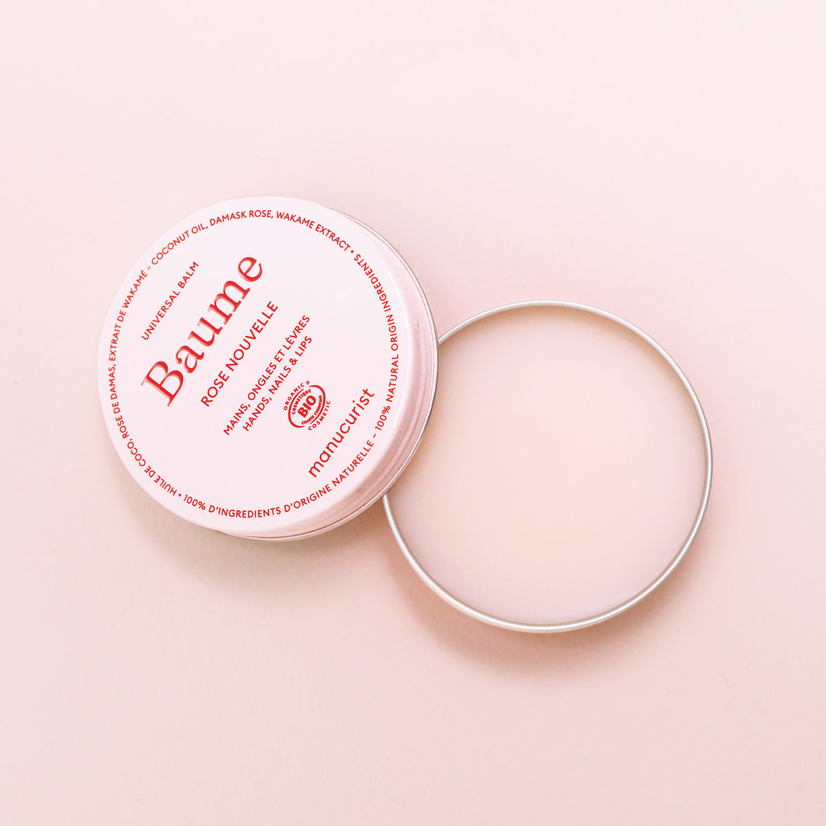 An open tin of Manucurist Natural Nourishing Balm - Cosmos Organic with a white lid and red text sits on a light pink surface. This multipurpose balm, smooth and unused, is ready to soothe and repair your skin.