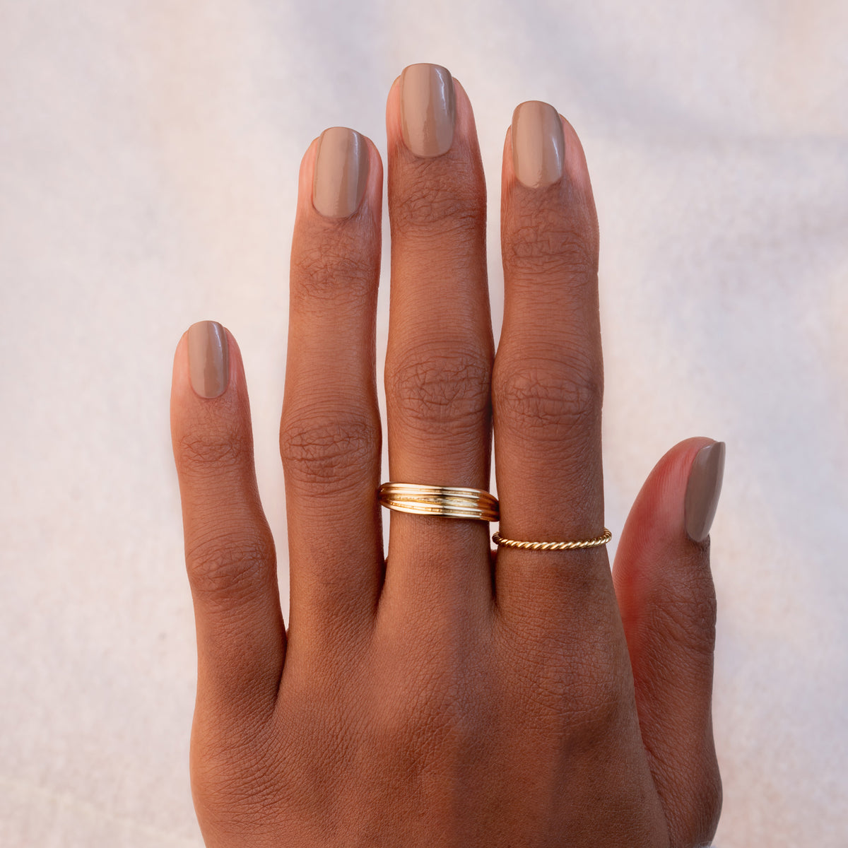 A hand with Manucurist Orme nail polish wears two gold rings—a thick band on the middle finger and a thin twisted band on the ring finger—set against a soft, neutral background.