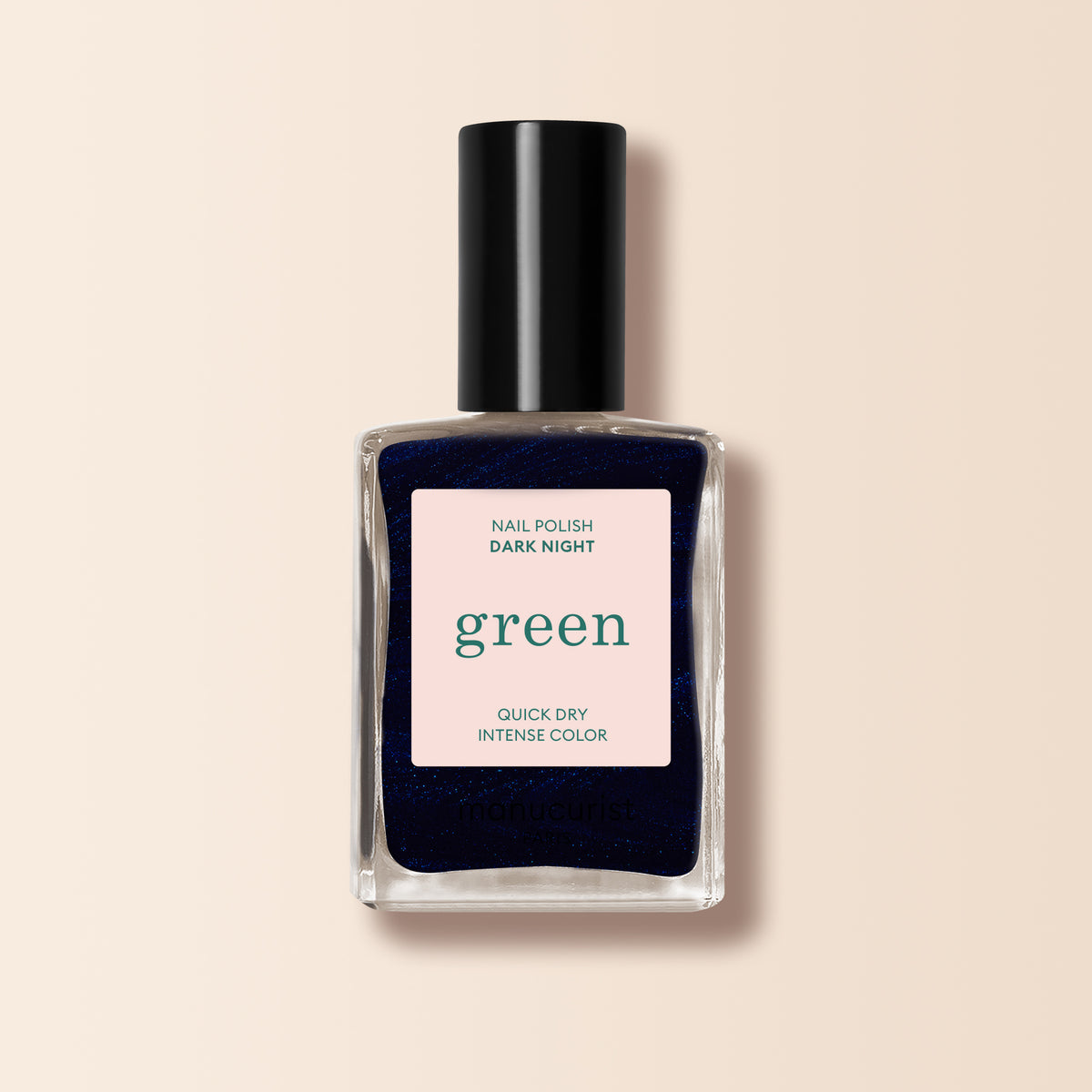 A bottle of Manucurist Dark Night nail polish with a black cap and green "Quick Dry Intense Color" label stands against a light beige background. The polish inside is a deep, nearly black midnight blue shade.