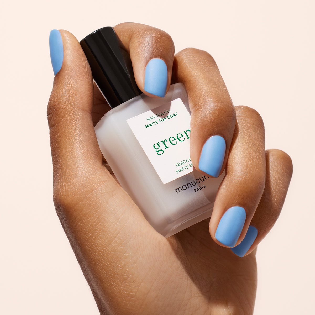 A hand with light blue matte nail polish holds a frosted bottle of Manucurist Matte Top Coat with a black cap against a light background.