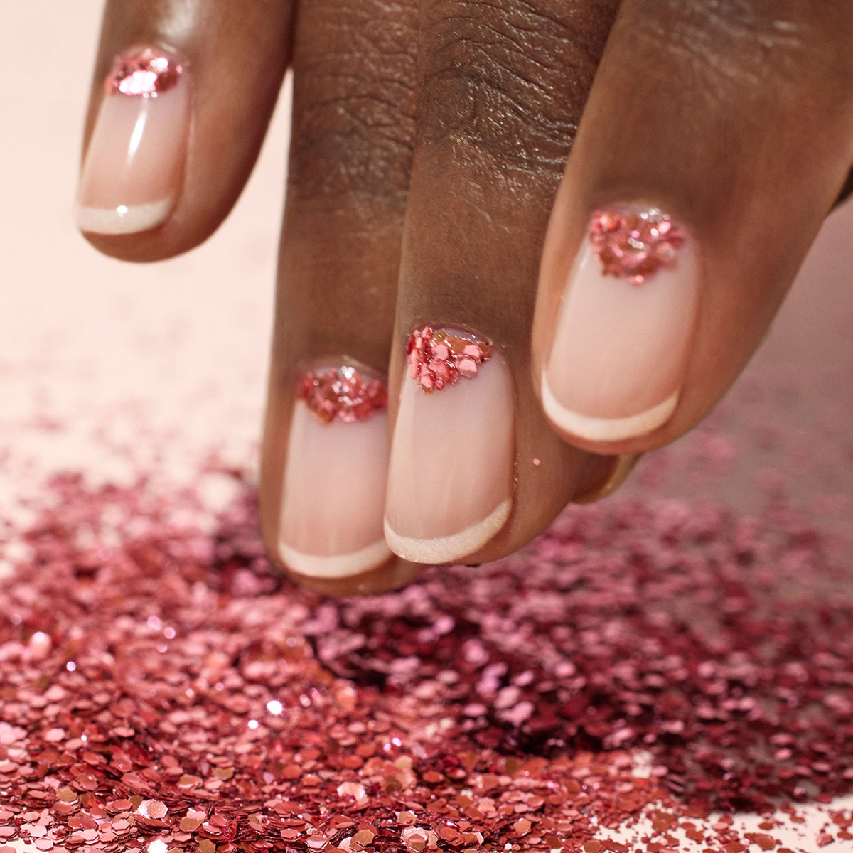 A hand with nude French tip gel nails adorned with pink glitter hearts near the cuticles rests above a surface sprinkled with Manucurist&