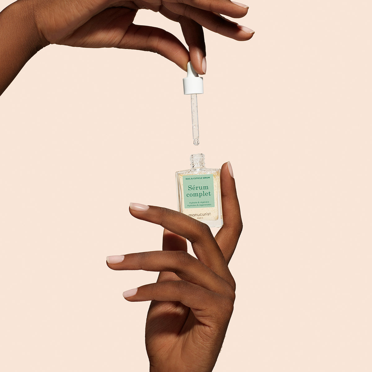 A person holds a Manucurist + Nail Hydrating Serum glass bottle in one hand and removes the dropper with the other, showing clear nail treatment drops in the dropper against a beige background.