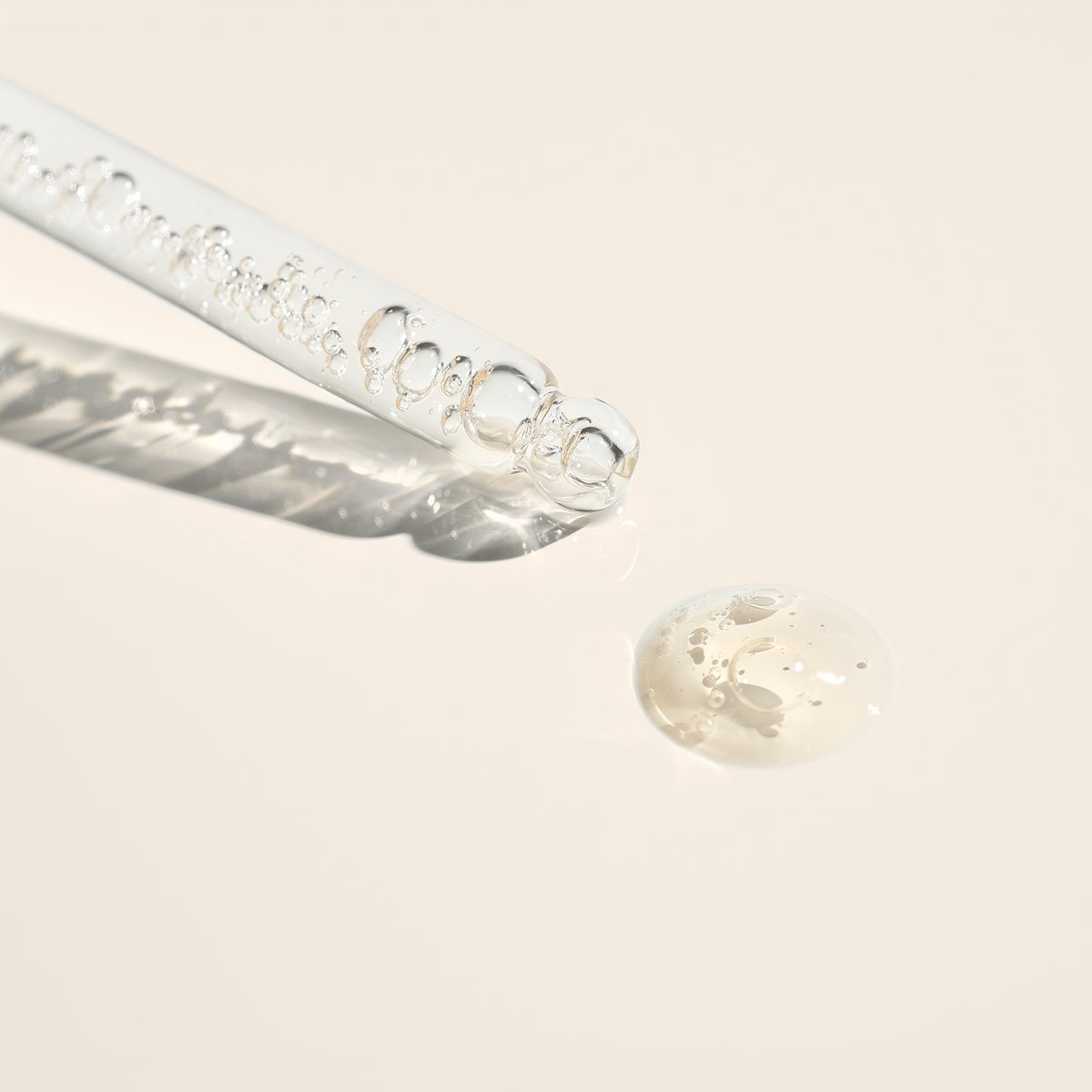 A close-up of the Manucurist Complete Serum glass dropper with clear liquid and bubbles, resting next to a small droplet of hydrating serum on a light beige surface.
