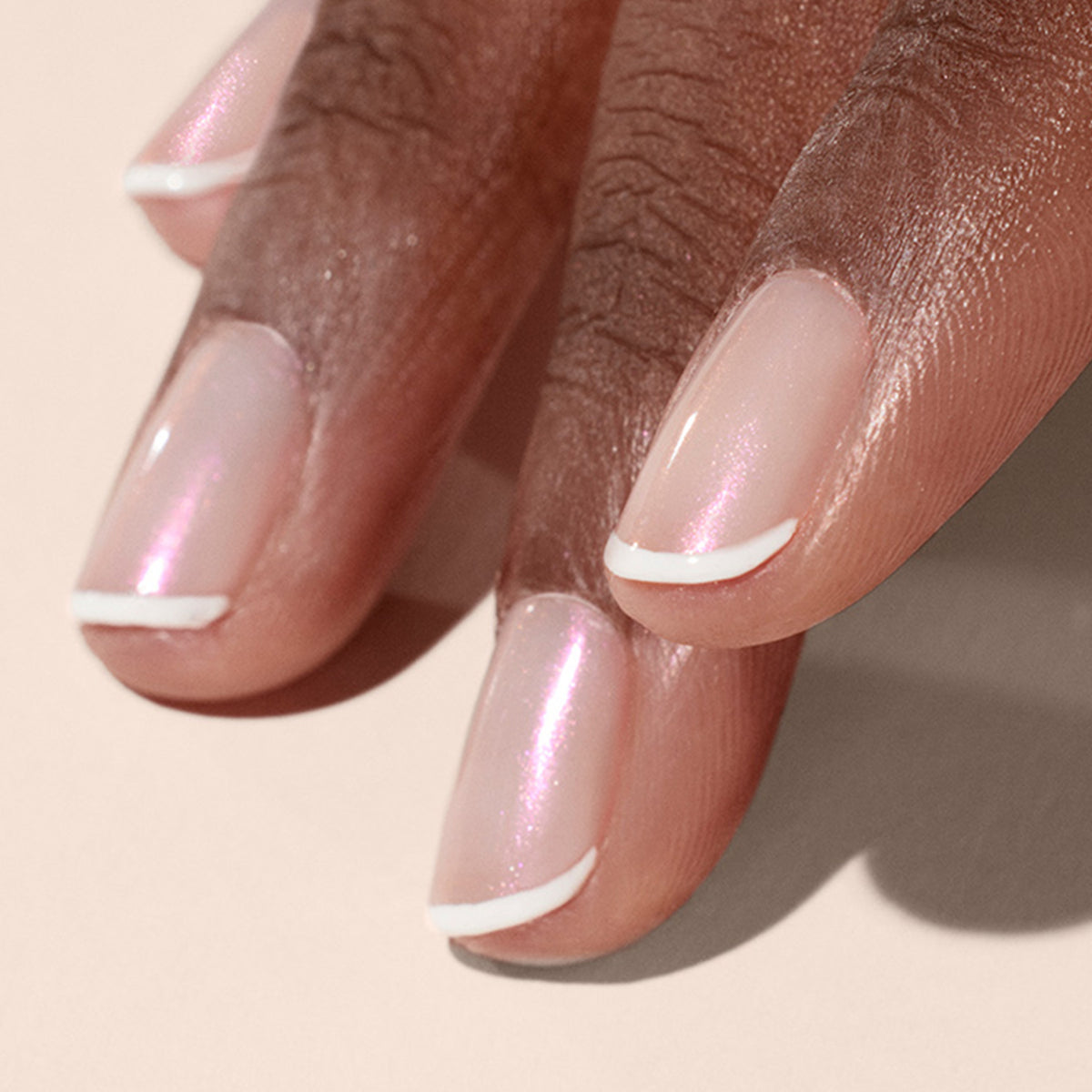 A close-up of a hand with medium-dark skin tone, nails painted in Manucurist’s Pink Glazed Effect Powder for a pearlescent pale pink look with thin white French tips. The background is soft beige.