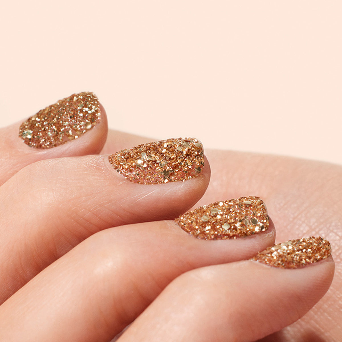 A close-up shows a hand with nails painted in Manucurist Rose Gold biodegradable glitter, creating elegant nail art against a soft beige background.