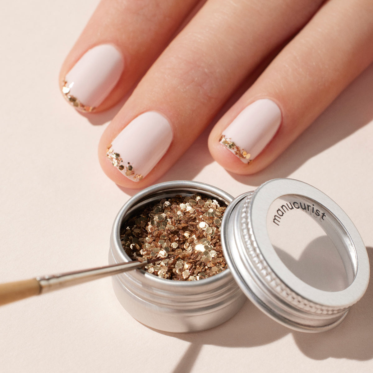A hand with neatly manicured nails features pale pink polish and rose gold glitter tips using Manucurist Rose Gold biodegradable glitter. An open container and a brush sit nearby, ideal for creating elegant nail art.