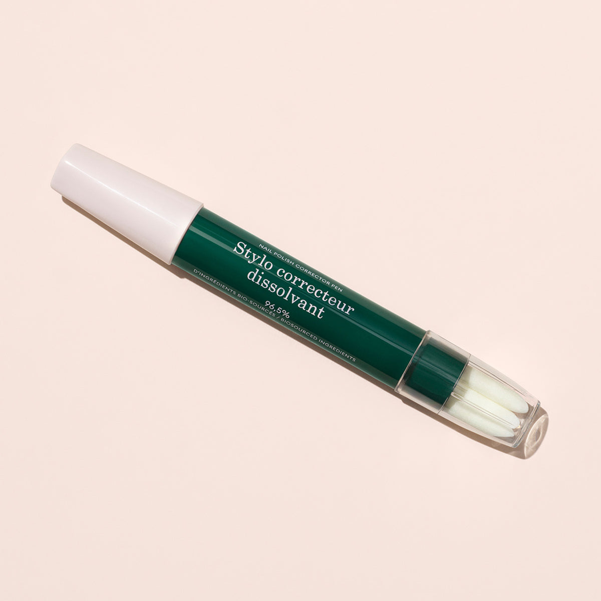 The Nail Polish Corrector Pen, featuring a green and white design with "Stylo correcteur dissolvant" printed on it and a clear cap revealing a white brush tip, lies on a light beige surface.