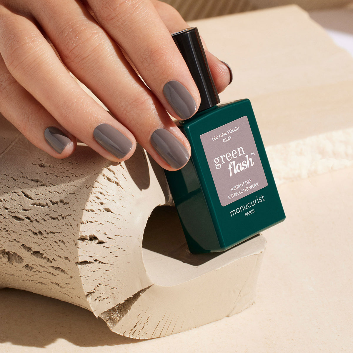 A hand with short, neatly manicured nails painted in glossy taupe gel polish holds a green bottle of Manucurist Clay gel polish against a textured, beige background.