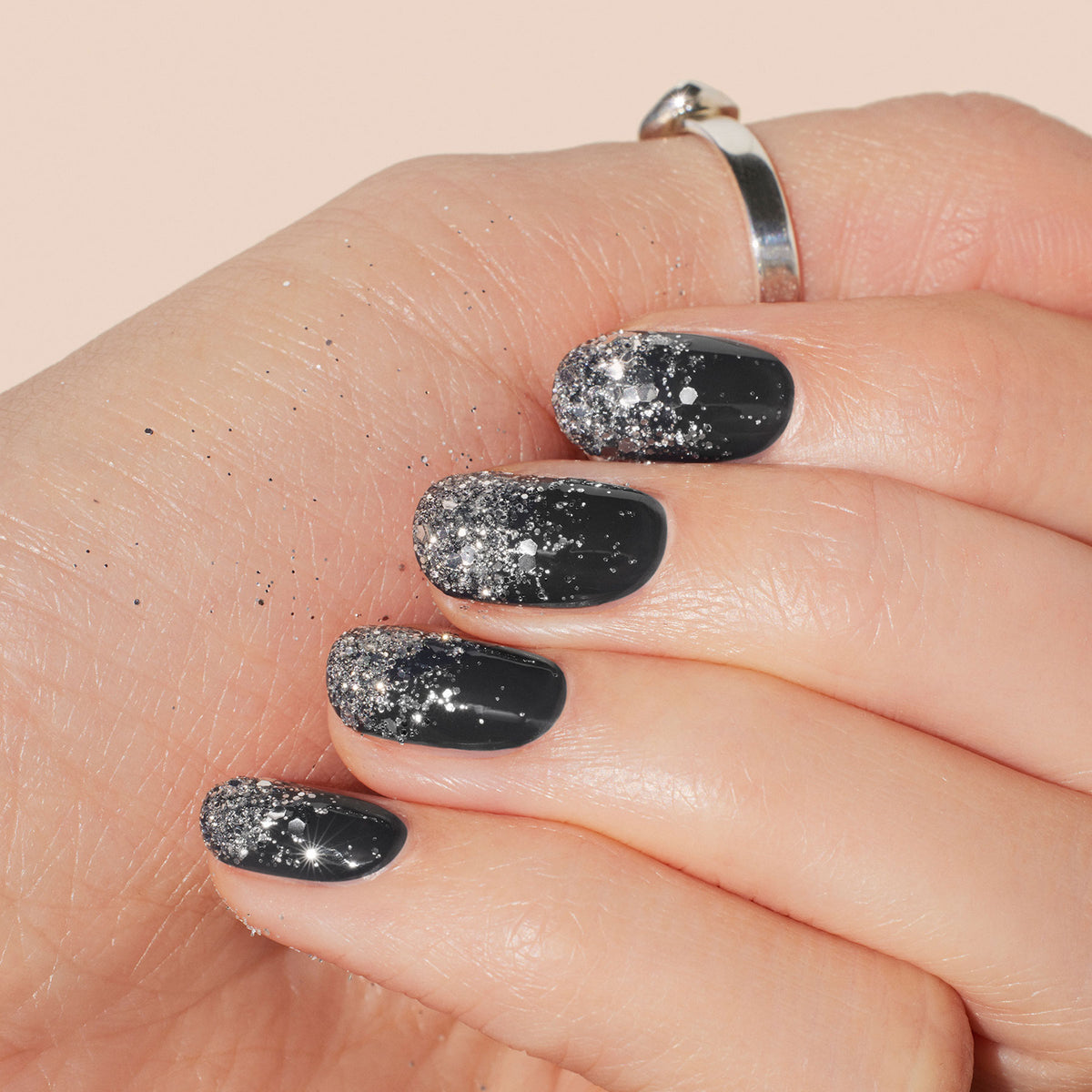 A close-up of a hand with short, oval nails painted glossy black and topped with Manucurist Silver biodegradable glitter at the tips. A simple silver ring adorns the middle finger.