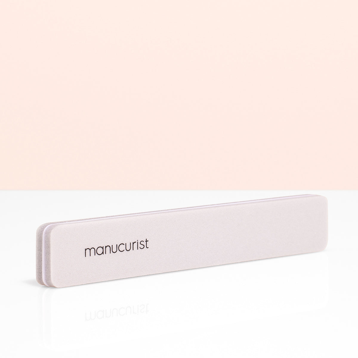 The Manucurist Nail buffer sponge in light pink, featuring the brand name, sits on a white reflective surface with a peach background—perfect for prepping your nail plate and improving polish staying power.