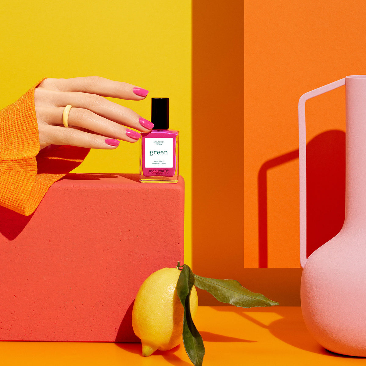 A hand with pink nails holds a Manucurist Pétula perfume bottle against a coral block, with a lemon and pink vase nearby, all set against a vibrant orange and yellow background.