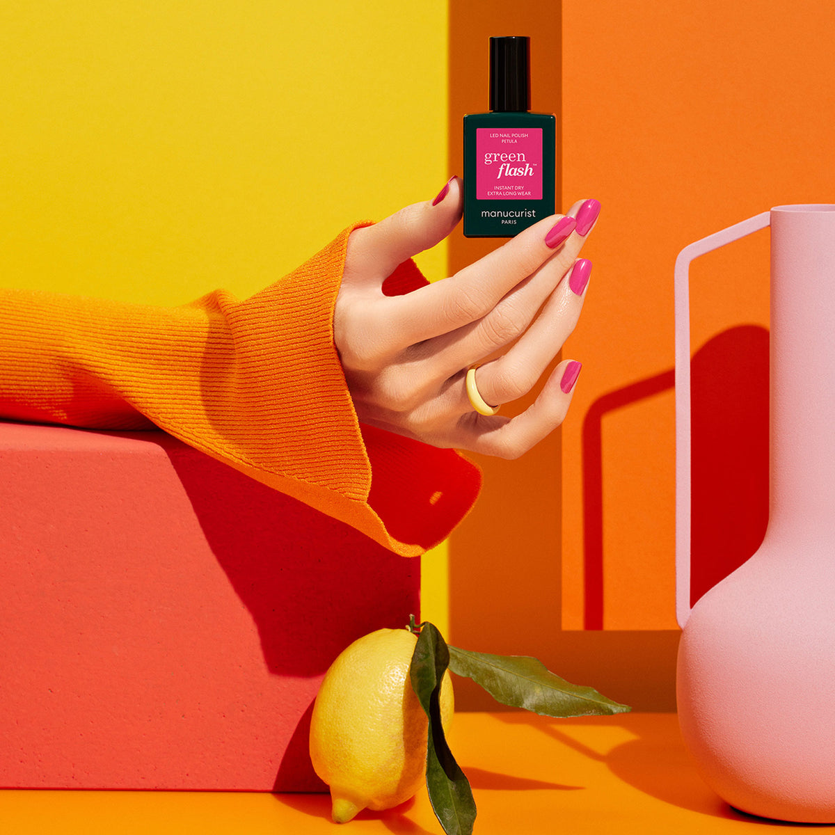 A hand with pink gel polish holds a bottle of Manucurist Pétula against a vivid orange and yellow background, with a lemon and pink pitcher nearby. An orange textured sleeve highlights this perfect summer shade.