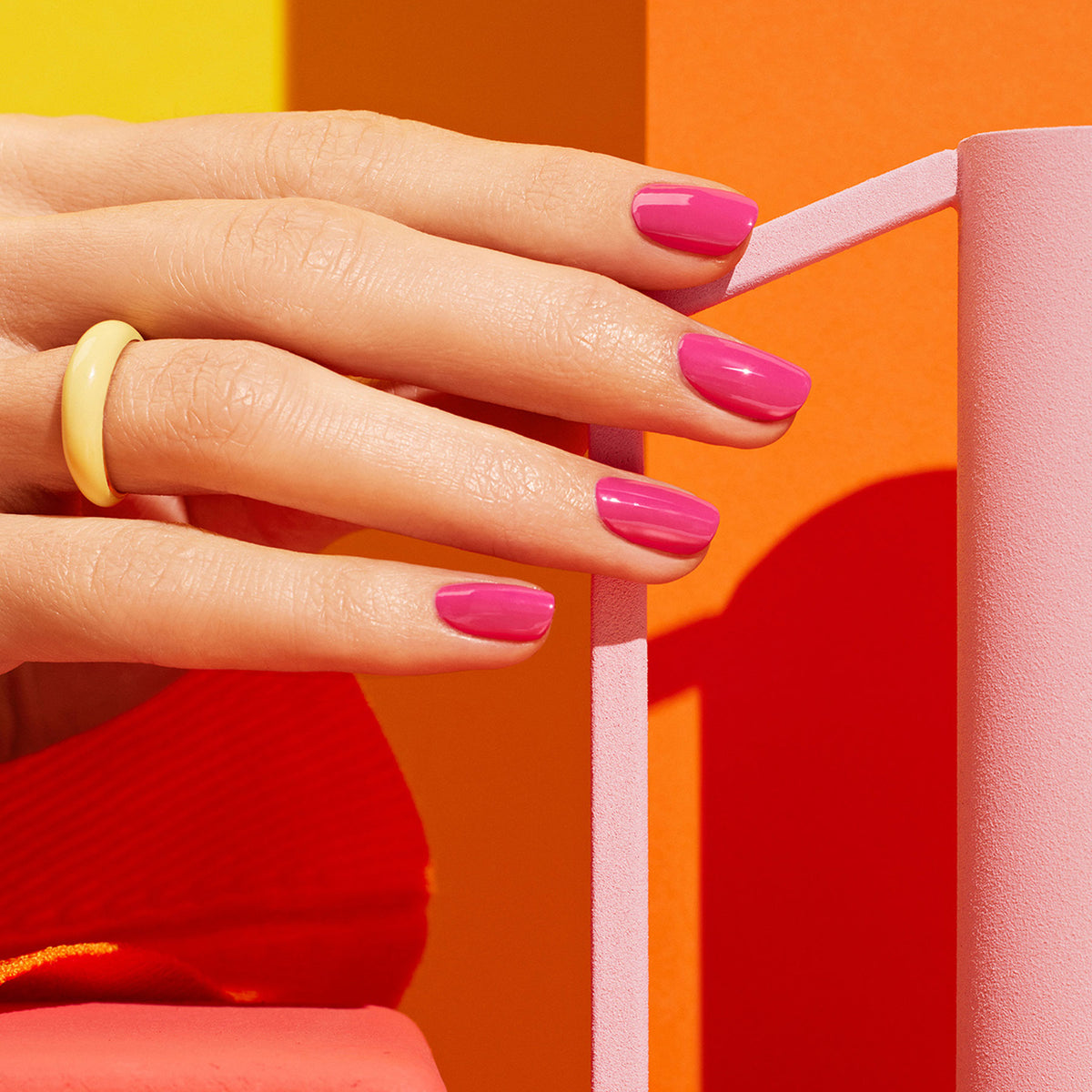 A hand with glossy Pétula pink nail polish by Manucurist rests on a geometric pink structure against a bold orange and yellow backdrop, accented by a chunky yellow ring on the index finger.