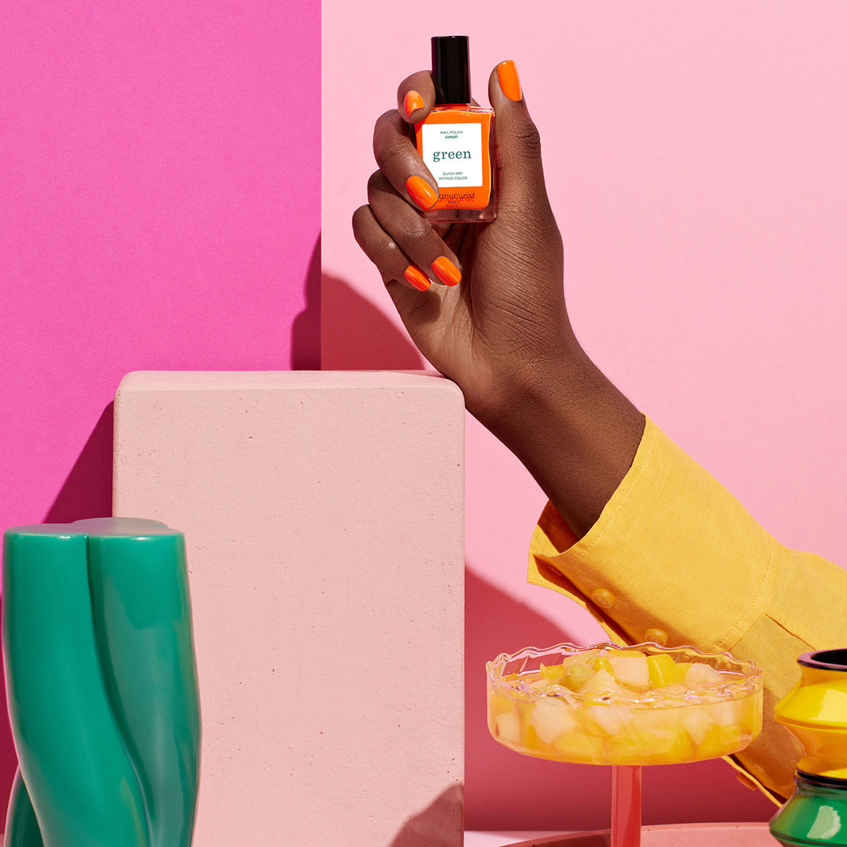 A hand with vibrant Sunset by Manucurist orange-painted nails holds the matching bottle against a pink background. The person wears a yellow sleeve. Beside them: a Green™ ceramic vase and a glass dish with ice and citrus pieces.