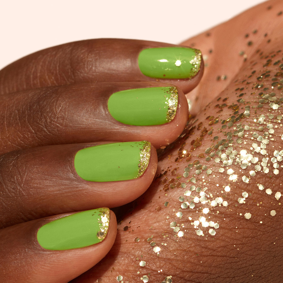 A hand with green nail polish and gold tips rests on skin sprinkled with Manucurist Gold biodegradable glitter.